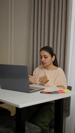 Vertical-video-of-a-happy-brunette-girl-in-a-beige-sweater-sitting-in-front-of-a-gray-laptop-and-communicating-via-video-with-a-teacher-while-studying-a-foreign-language-German-in-a-modern-apartment-during-the-day